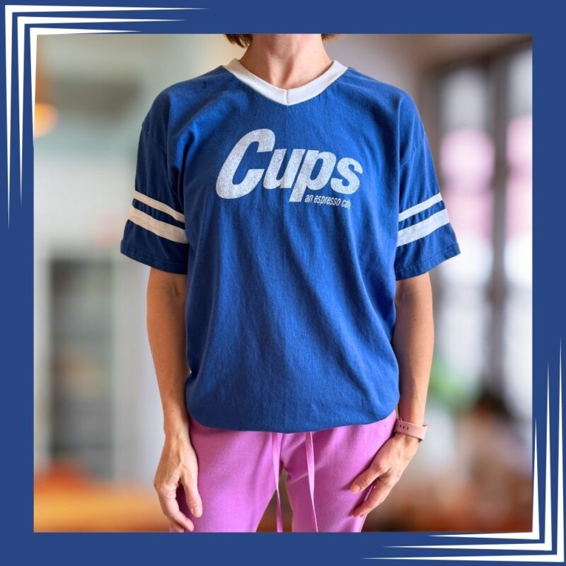 Front view of blue Cups logo V-neck jersey t-shirt with white athletic stripes on sleeves, modeled with pink pants against a cafe background.